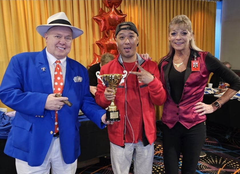Three people pose together at a casino event, with the person in the center holding a trophy and flashing a peace sign, surrounded by red star balloons and gold curtains.